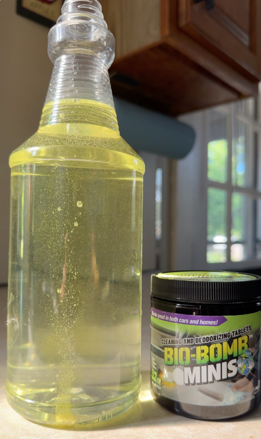 A clear plastic spray bottle filled with water and one Bio-Bombs Mini. Yellow-green cleaning liquid stands next to a black jar labeled Bio-Bomb Minis on a kitchen counter with cabinets and a window in the background.