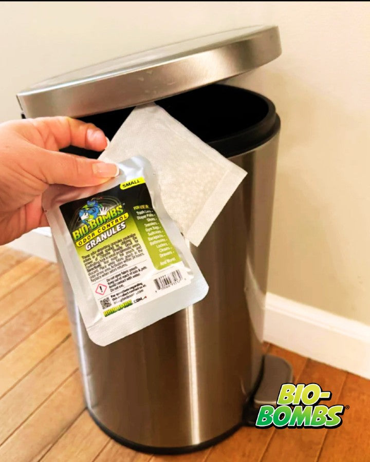 A hand holds a package of Bio-Bombs Odor Control Granules next to an open stainless steel trash can on a wooden floor.