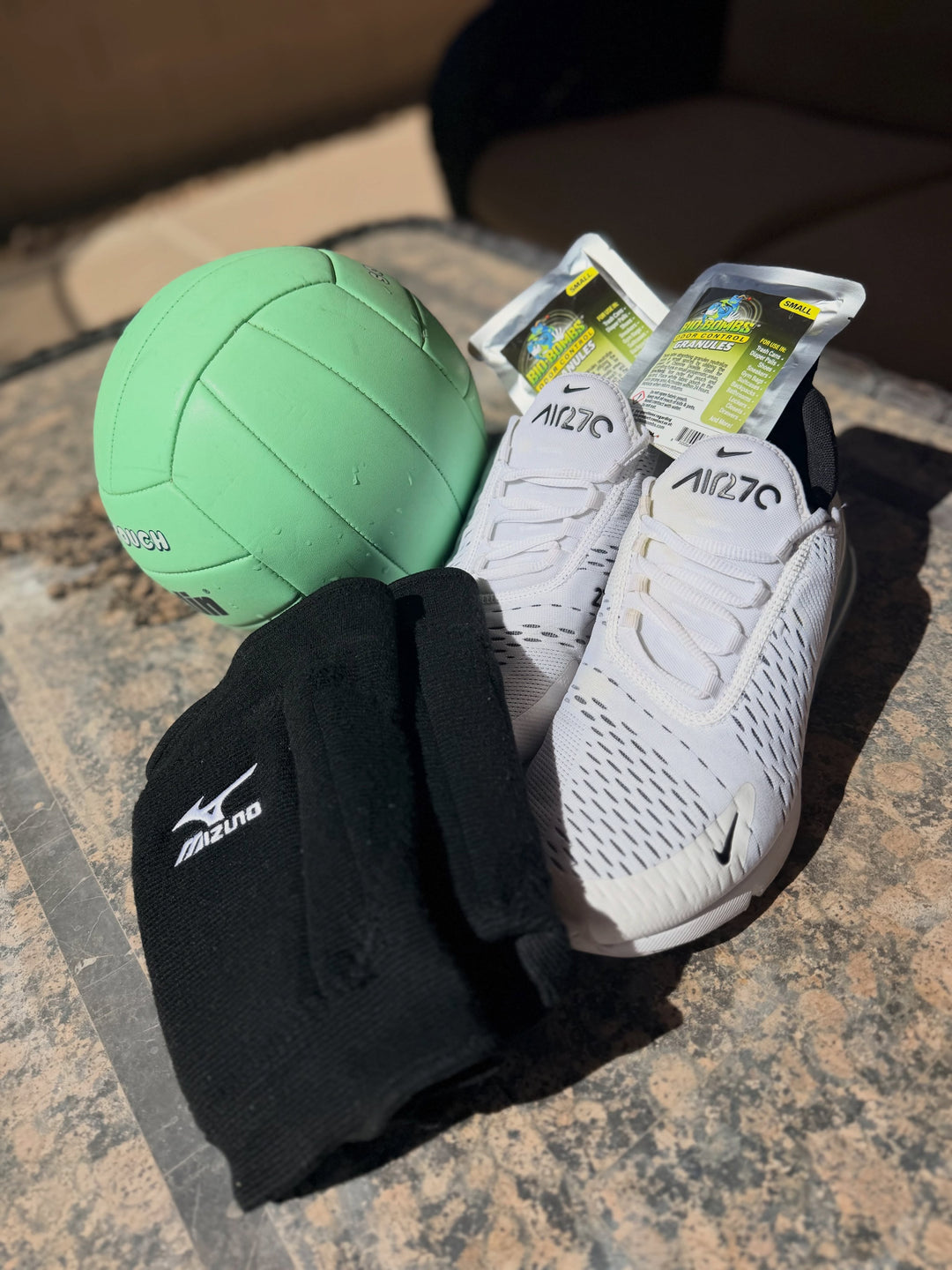 A green volleyball, a pair of white Mizuno running shoes, a black Mizuno knee pad, and two small odor control granules arranged on a stone surface.