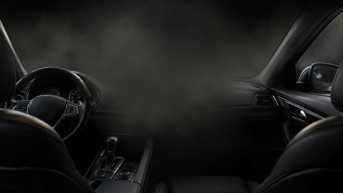 Dark, moody interior of a car with black leather seats, a steering wheel, gear shifter, and dashboard details, partially obscured by mist or smoke filling the center of the cabin.